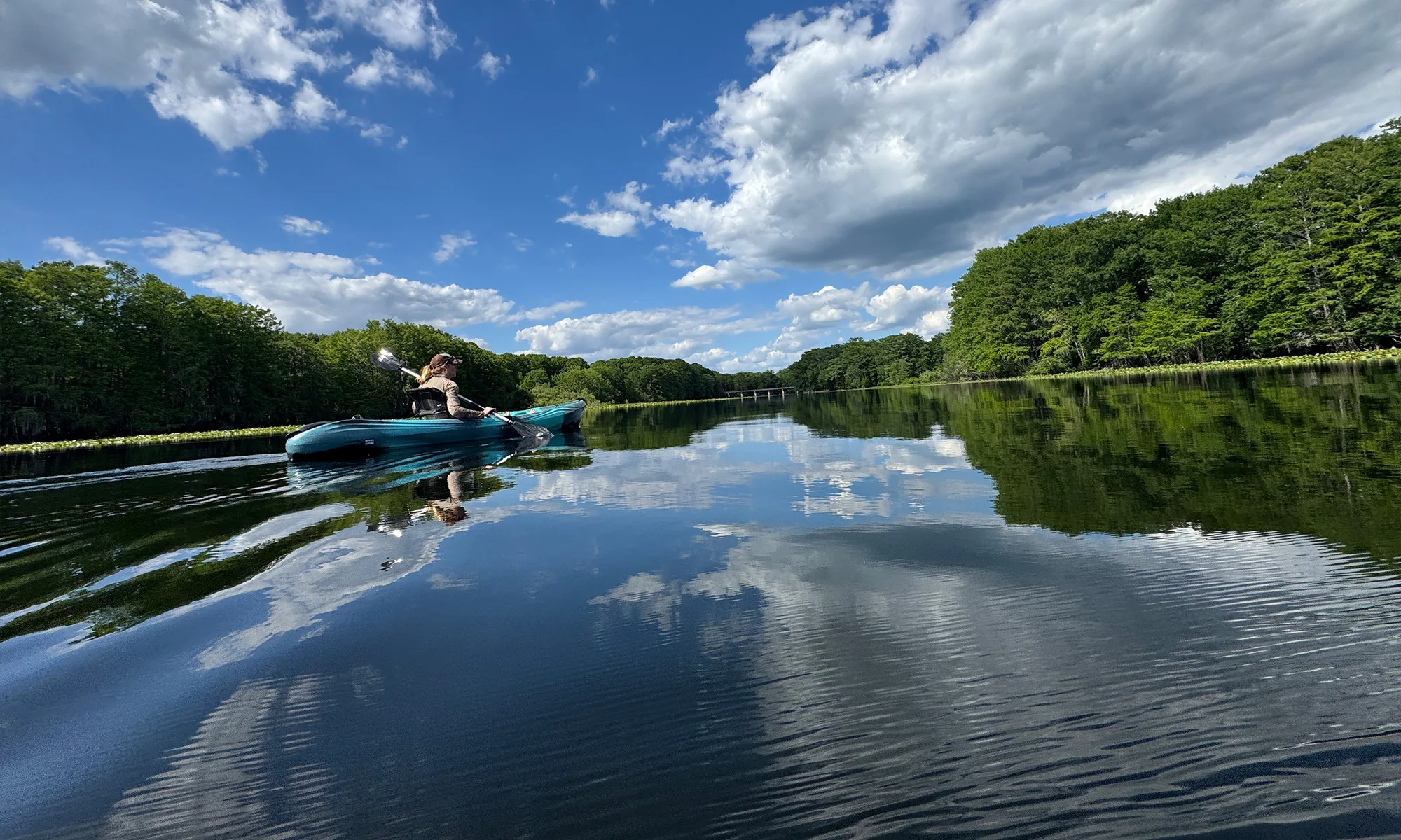 Person kayaking in a turquoise canoe on calm water with forested banks and a blue sky with clouds, reflections visible in the water