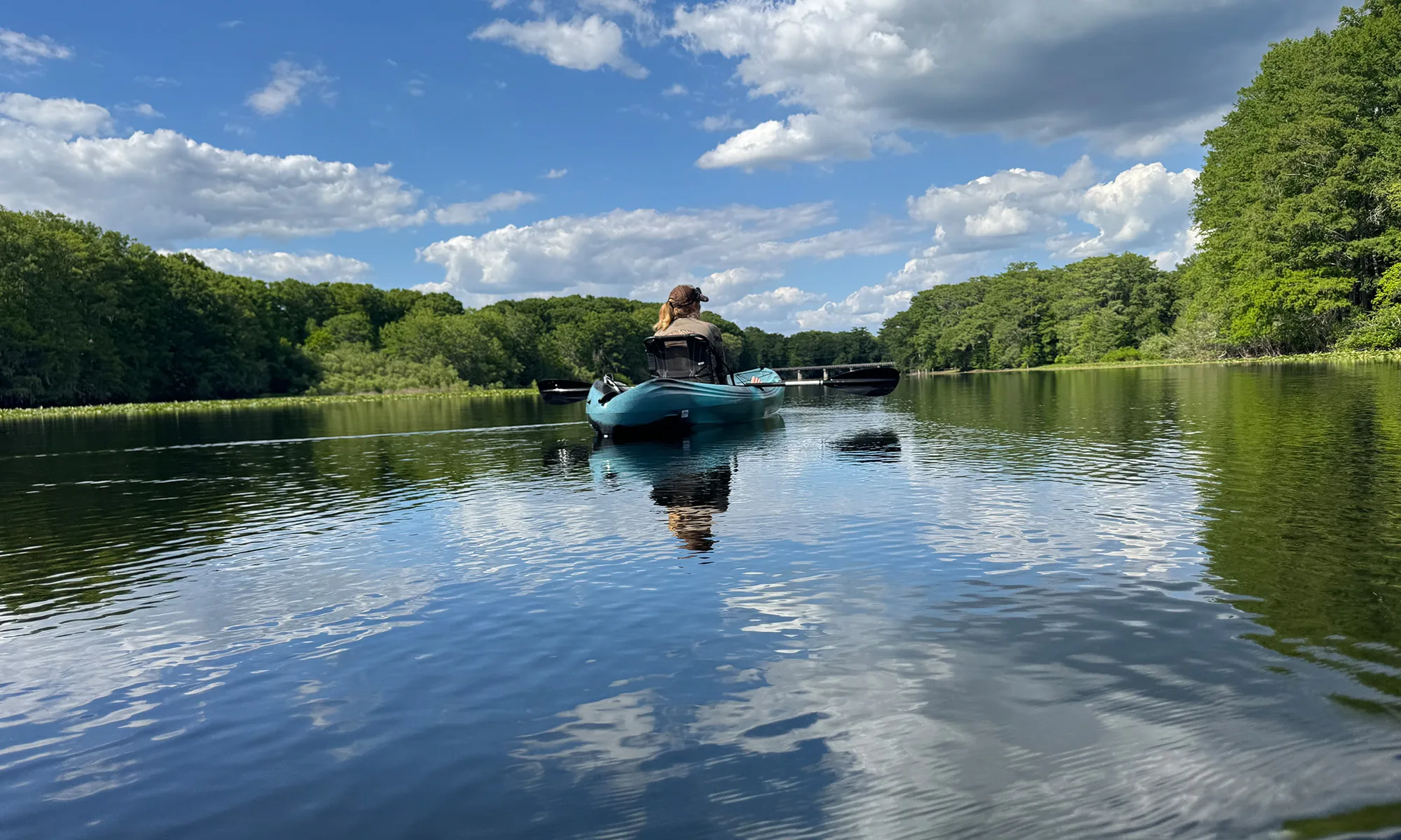 Person paddling a blue kayak on a calm lake, with green trees along the shoreline and a blue sky with white clouds above.