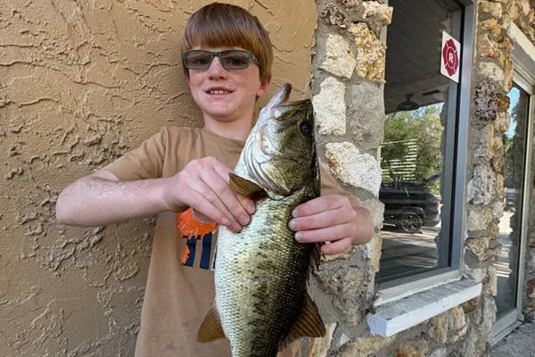 Boy wearing sunglasses holds a large bass up to camera beside a stone building wall. He smiles after a fishing catch.